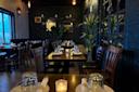 A dimly lit restaurant interior with wooden tables set for dining. Candles and glasses are placed on the tables. Dark green walls feature mirrors and light fixtures. Potted plants add a touch of greenery to the cozy atmosphere.