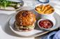 A gourmet burger topped with melted cheese and crispy onions on a brioche bun sits on a plate with a side of golden fries and a small bowl of ketchup. Blurred background includes grilled asparagus and greens on another dish.