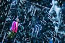 A person in a bright pink jacket stands amid digital projections of binary code and a large face, creating a futuristic, immersive atmosphere on stage.