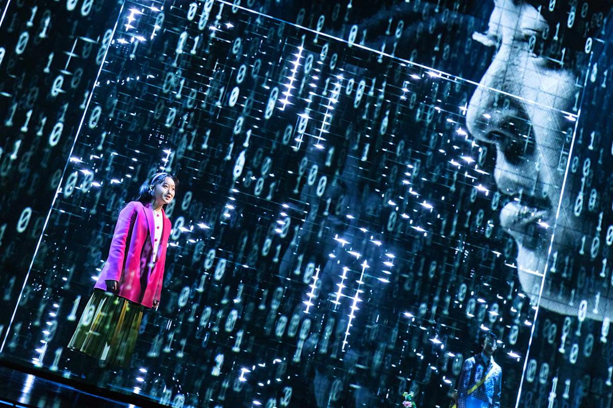 A person in a bright pink jacket stands amid digital projections of binary code and a large face, creating a futuristic, immersive atmosphere on stage.