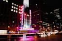 Night view of Radio City Music Hall in New York City, with bright neon lights and signs illuminating the building and reflecting on the wet street. A few people walk along the sidewalk.