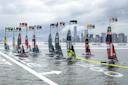 A group of high-tech sailboats race on the water near a city skyline, each boat displaying country flags and speed data above them. The image shows a competitive sailing event with various nations participating.