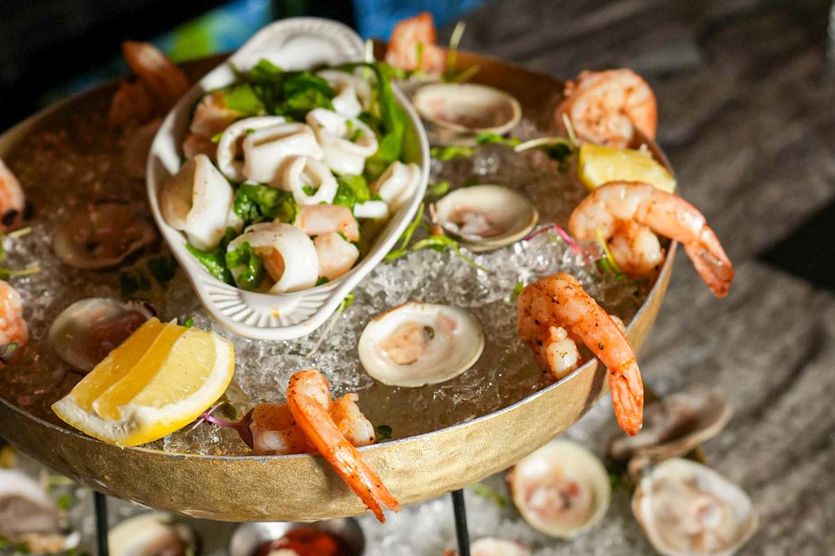 A seafood platter on ice, featuring shrimp, lemon wedges, clams, and a dish of mixed seafood salad garnished with greens.