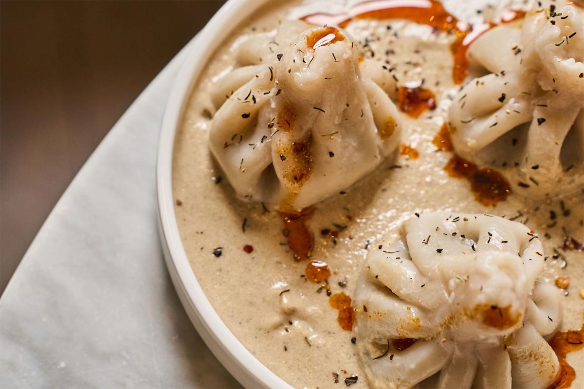Three steamed dumplings sit in a creamy beige sauce, garnished with herbs and drizzled with red chili oil, served on a white plate.