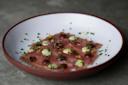 A plate of tuna crudo garnished with herbs, lemon zest, and drizzles of sauce. The tuna slices are arranged neatly on a white plate with a brown rim, creating a visually appealing dish on a gray surface.