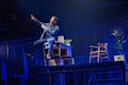 A performer in a denim jacket and jeans sits on the edge of a desk on stage, gesturing energetically. The set includes chairs, plants, and boxes, with a blue-lit cityscape backdrop and dramatic lighting.
