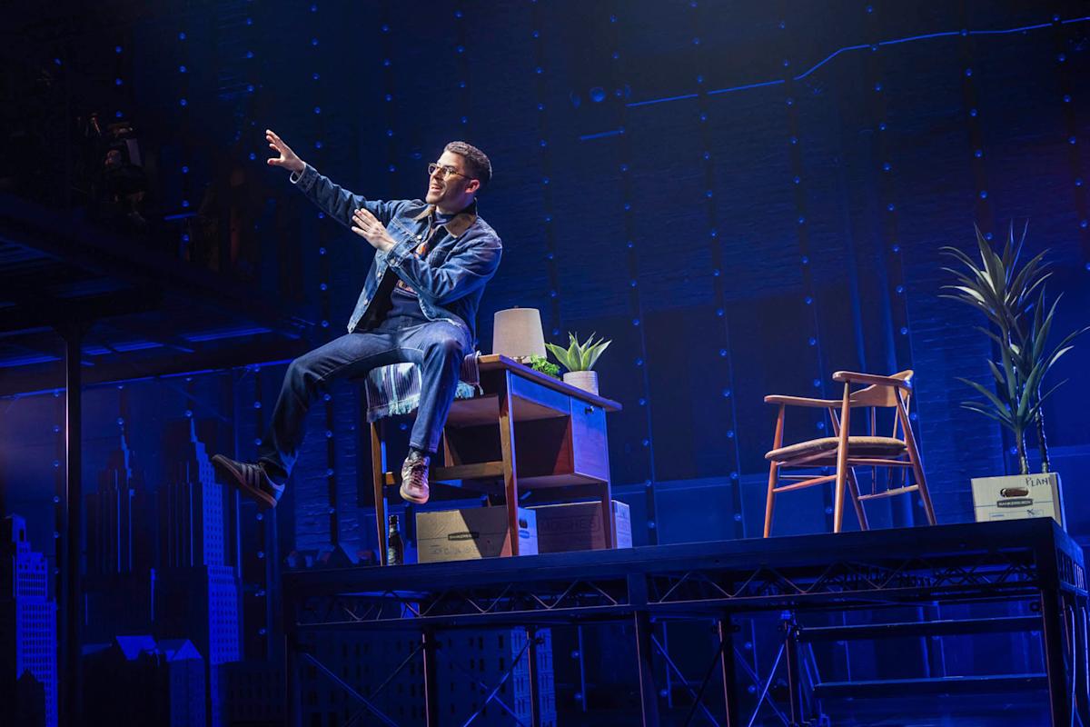 A performer in a denim jacket and jeans sits on the edge of a desk on stage, gesturing energetically. The set includes chairs, plants, and boxes, with a blue-lit cityscape backdrop and dramatic lighting.