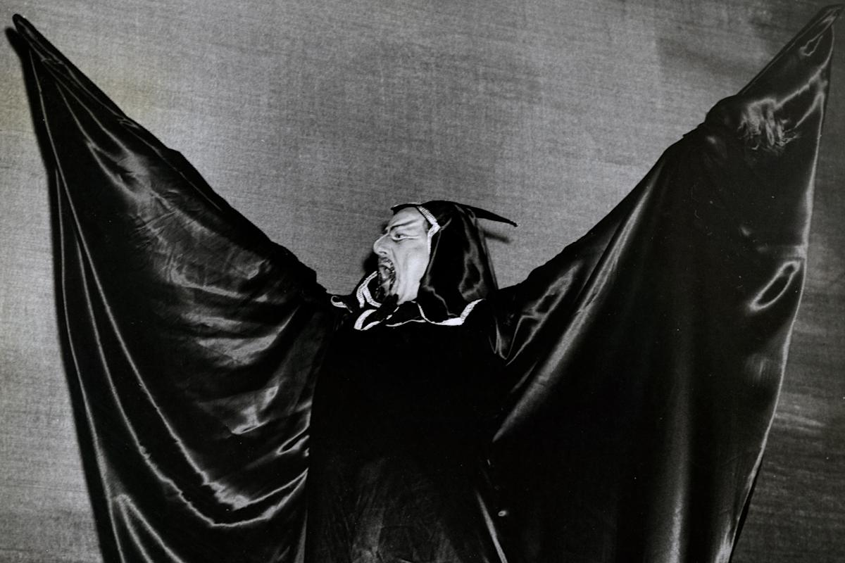 A person in a dramatic, dark costume spreads large, wing-like sleeves and wears a hood with small horns, posing theatrically against a plain background. The image is black and white.