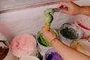A person scoops green ice cream into a cone from a tub, surrounded by other colorful ice cream tubs in a freezer.
