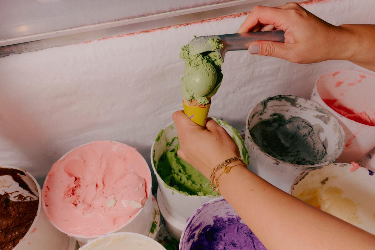 A person scoops green ice cream into a cone from a tub, surrounded by other colorful ice cream tubs in a freezer.
