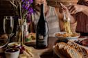 Two people dine at a table set with pasta, wine, bread, cheese, grapes, and flowers. One person uses utensils to serve pasta, while the other sits nearby. A bottle of WillaKenzie Estate wine is in focus in the center.