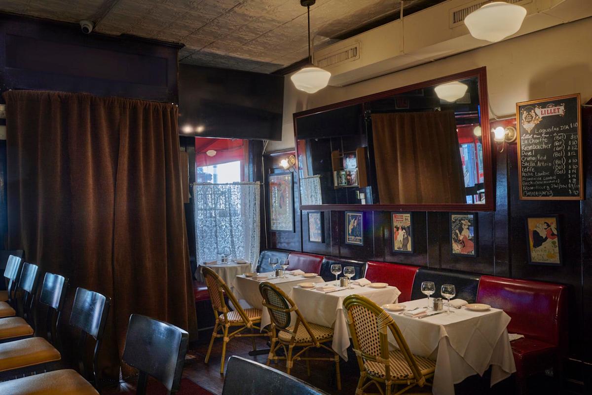 A cozy restaurant interior with tables set for dining. Tables have white tablecloths, and wine glasses are arranged neatly. Red booth seating and wicker chairs are present. Walls are decorated with mirrors and vintage posters. Soft lighting adds warmth.