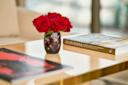 A glass vase with red roses sits on a wooden table next to two books, one titled "Alan Faena," with a blurred, modern background.
