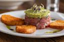 A plate with tuna tartare topped with avocado and microgreens, surrounded by three toasted bread slices and decorative drizzles of sauce.