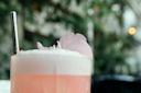 A close-up of a pink creamy beverage garnished with a flower petal, served in a glass with a metal straw. The background is softly blurred with hints of greenery, creating a serene and refreshing atmosphere.
