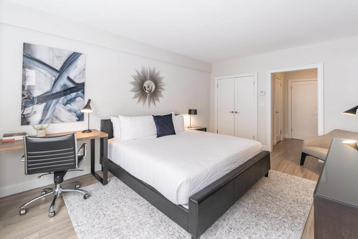 Modern bedroom with a large bed, white bedding, and a black frame. A sleek desk and chair are on the left, and a sunburst mirror hangs above the bed. There's a contemporary abstract painting on the wall and ambient lighting throughout.