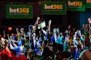 A large, energetic crowd cheers and raises their arms at an indoor sports event, with prominent green “bet365” banners hanging in the background. The atmosphere is festive and lively.