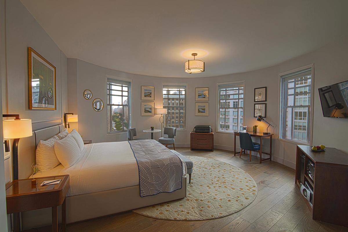 A spacious hotel room with a large bed, two nightstands, a round rug, a desk with a chair, a TV on the wall, and several windows providing natural light and views of city buildings outside.
