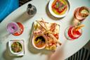 A marble table with various drinks, a small plate of green olives, and a charcuterie board featuring cured meats, cheese, breadsticks, and spreads, photographed from above in a bright setting.