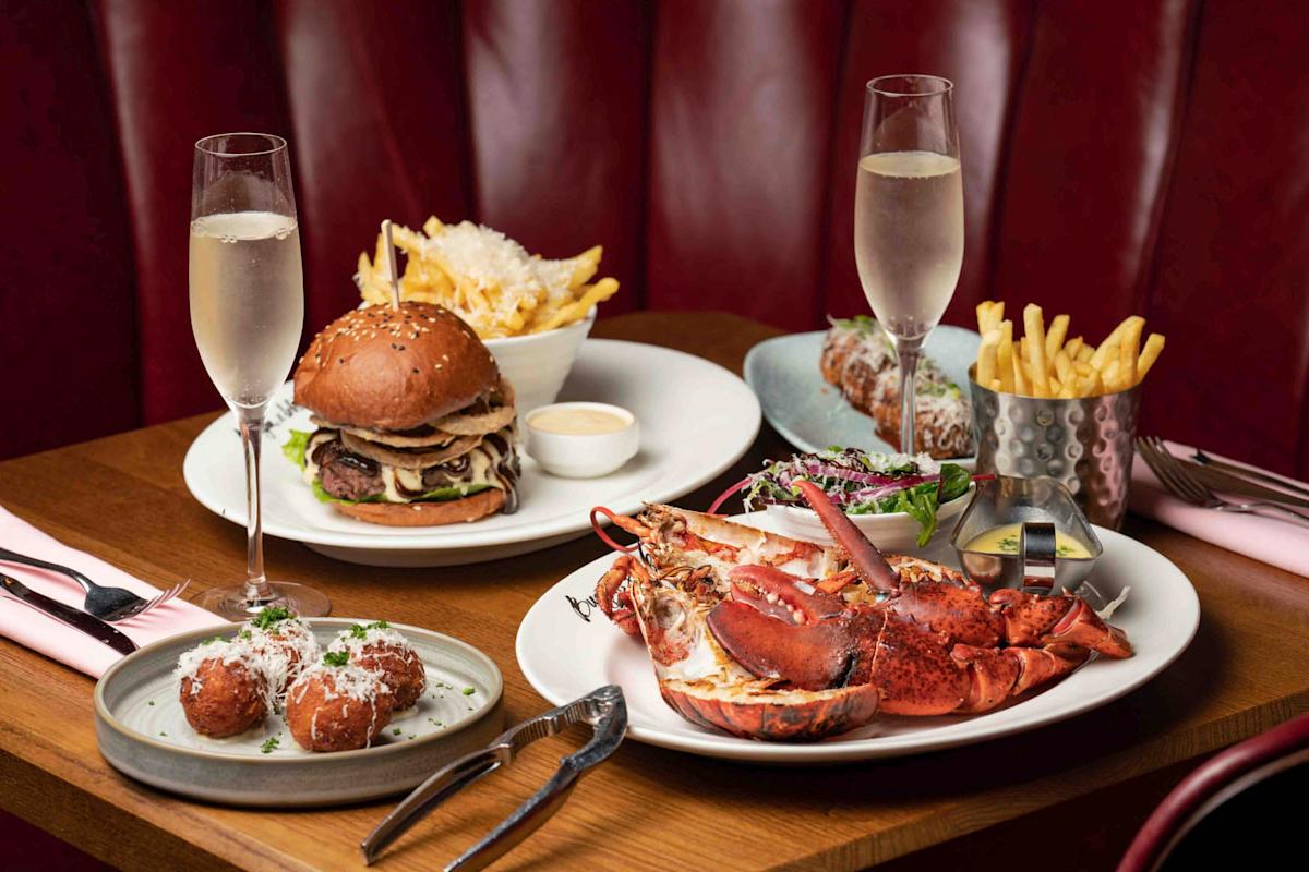 A table with a gourmet spread: lobster and crab on a plate, truffle fries in a metal cup, a loaded burger with fries, arancini balls, and two glasses of sparkling wine, all set against a red leather booth backdrop.