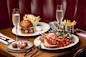 A table with a gourmet spread: lobster and crab on a plate, truffle fries in a metal cup, a loaded burger with fries, arancini balls, and two glasses of sparkling wine, all set against a red leather booth backdrop.