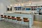 A modern diner interior featuring a marble counter with brown stools, overhead menu boards listing items like Caviar & Champagne, Potato Latkes, and Egg Creams. The counter displays various syrups, and a drink dispenser is on the right. 