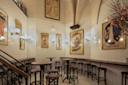A vintage-style room with white tiled walls and art nouveau posters. The room features several tall, round tables and wooden stools. Ornate lighting fixtures hang on the walls, and a staircase with a black railing is visible on the left.