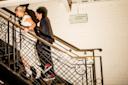Two people walk up a staircase with decorative railing in a well-lit area. One wears a white dress, the other wears dark pants and jacket. A sign on the white brick wall reads "COMMISSARY BALCONY.