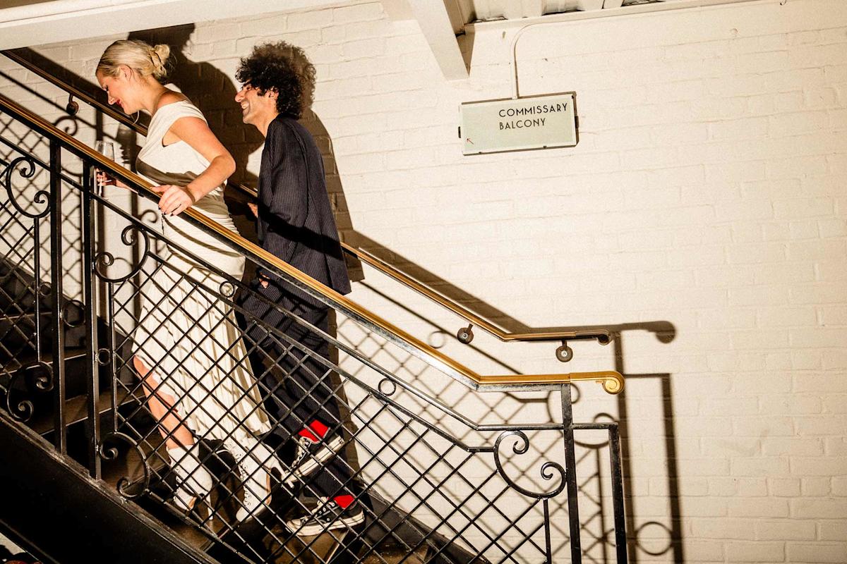 Two people walk up a staircase with decorative railing in a well-lit area. One wears a white dress, the other wears dark pants and jacket. A sign on the white brick wall reads "COMMISSARY BALCONY.