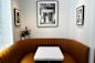 A cozy corner booth with a brown leather banquette and a white square table. Black and white framed photos decorate the white walls, and a small plant adds a touch of greenery.