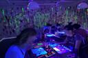 People painting with glowing neon colors under blacklights in a dark room, with splattered and doodled neon artwork on the walls. Paintings and art supplies are illuminated on the tables as they work.