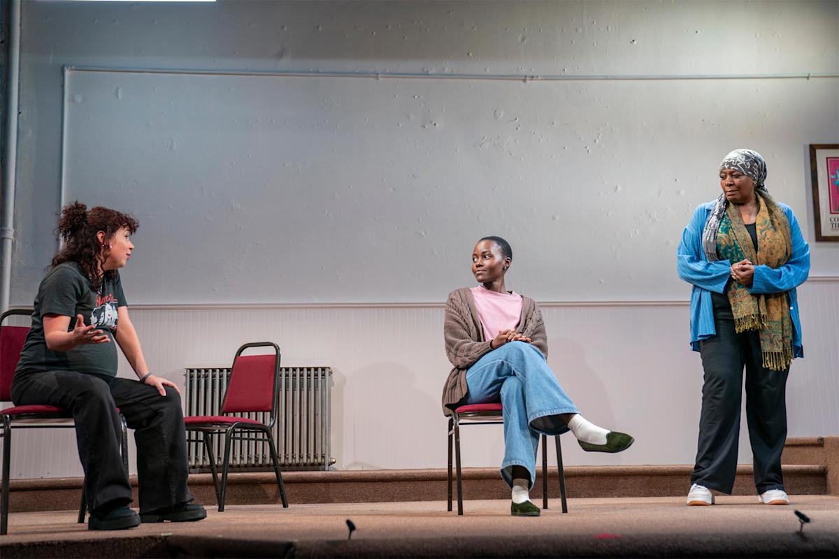 Three people sit on chairs on a stage, engaged in conversation. One gestures while talking, another listens with arms crossed, and the third stands facing them. The stage has minimal props and a plain background.