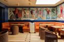 A stylish restaurant interior with orange upholstered seating, wicker chairs, and wooden tables. A colorful mural depicting people and animals in a jungle scene decorates the wall above the banquettes.