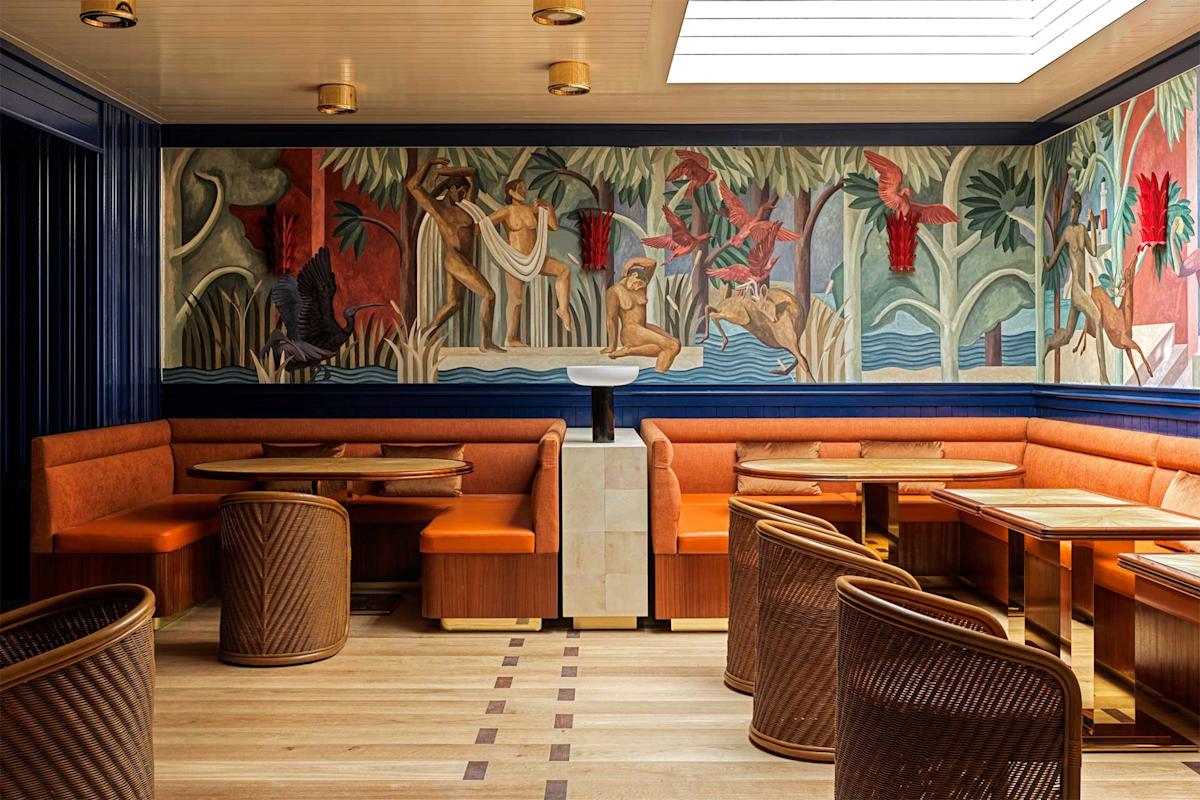 A stylish restaurant interior with orange upholstered seating, wicker chairs, and wooden tables. A colorful mural depicting people and animals in a jungle scene decorates the wall above the banquettes.