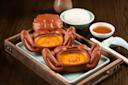Ceramic crab-shaped bowls filled with a creamy orange dish are placed on a tray. A bowl of white rice, a small dish of sauce, and a ceramic spoon with chopsticks are placed nearby on the wooden table.