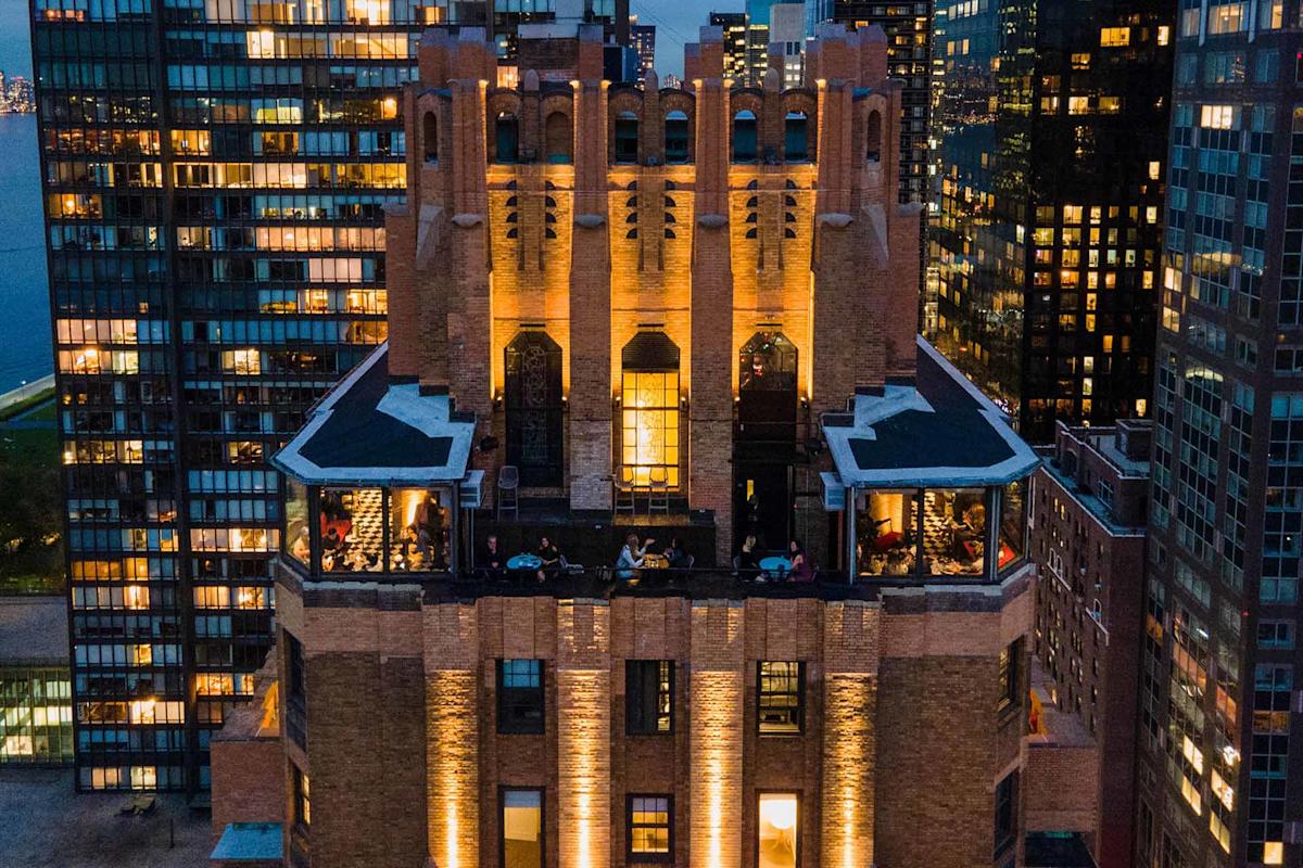 Aerial view of a tall, illuminated brick building with gothic architectural details, surrounded by modern glass skyscrapers at dusk. People are visible dining on terraces. City lights glow in the background.