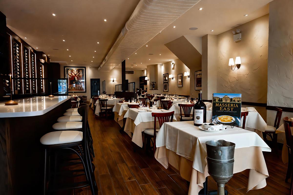 Elegant restaurant with white tablecloths, wooden chairs, a bar with high stools, and wine bottles on display. Warm lighting creates a cozy ambiance. A large menu and a plate of pasta sit on a table in the foreground.