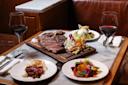 A table set for two with glasses of red wine, a wooden board with grilled steak and salt, and plates of salad, vegetables, and a meat appetizer in a cozy restaurant booth.