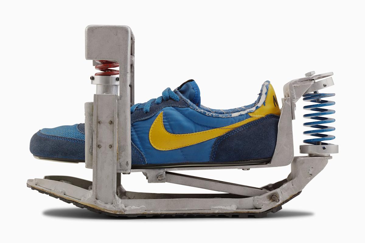A blue and yellow Nike sneaker is mounted in a metal contraption with red and blue springs, resembling an experimental device for testing or enhancing the shoe’s movement.
