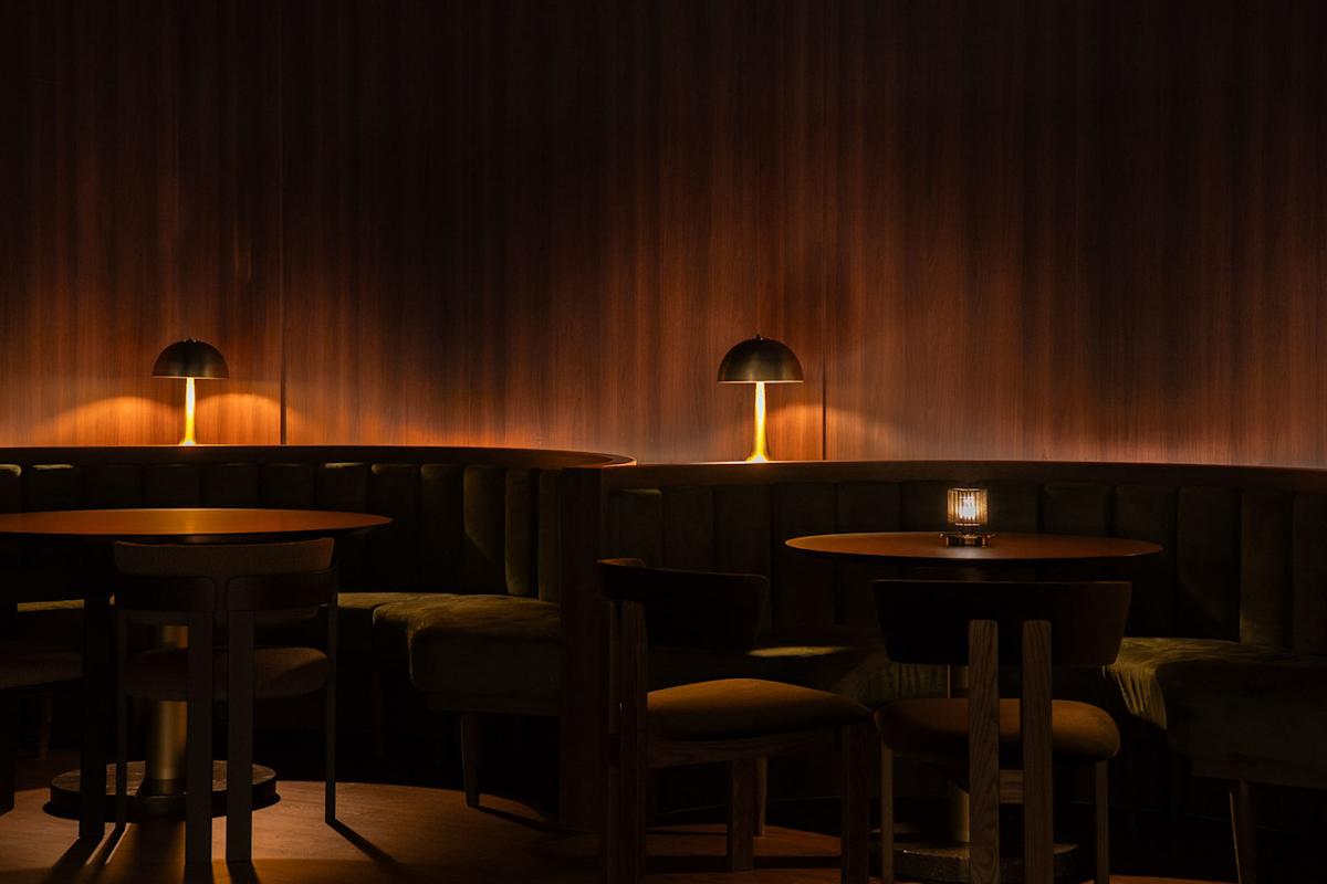 Dimly lit lounge with round wooden tables, curved velvet banquettes, and warm light from modern table lamps against a dark wooden wall.