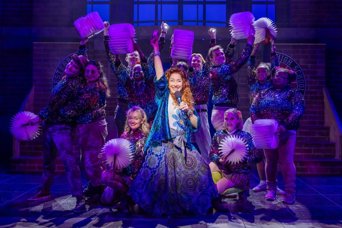 A woman sings joyfully on stage, surrounded by dancers holding white lanterns and wearing sparkly tops. The group poses energetically under purple stage lights, with a brick school backdrop behind them.