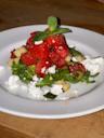 A salad featuring chunks of watermelon, cucumber, arugula, and crumbled feta cheese, garnished with a fresh mint sprig, served on a white plate atop a wooden surface.