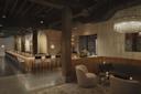 Dimly lit, modern bar interior with a long counter lined with high stools on the left. A crystal chandelier hangs over a cozy seating area with two round chairs and a tufted sofa on the right. Dark wooden beams and shelves add warmth to the space.