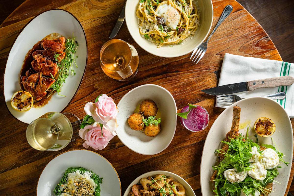 A wooden table set with various dishes, including pasta, fried arancini, salad, roasted meat, and two glasses of wine, alongside pink flowers and silverware on a napkin.