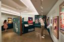 A museum exhibit features vintage wildlife protection and history posters. Two people observe displays on teal walls under bright lighting. The polished concrete floor reflects the vibrant, colorful posters.