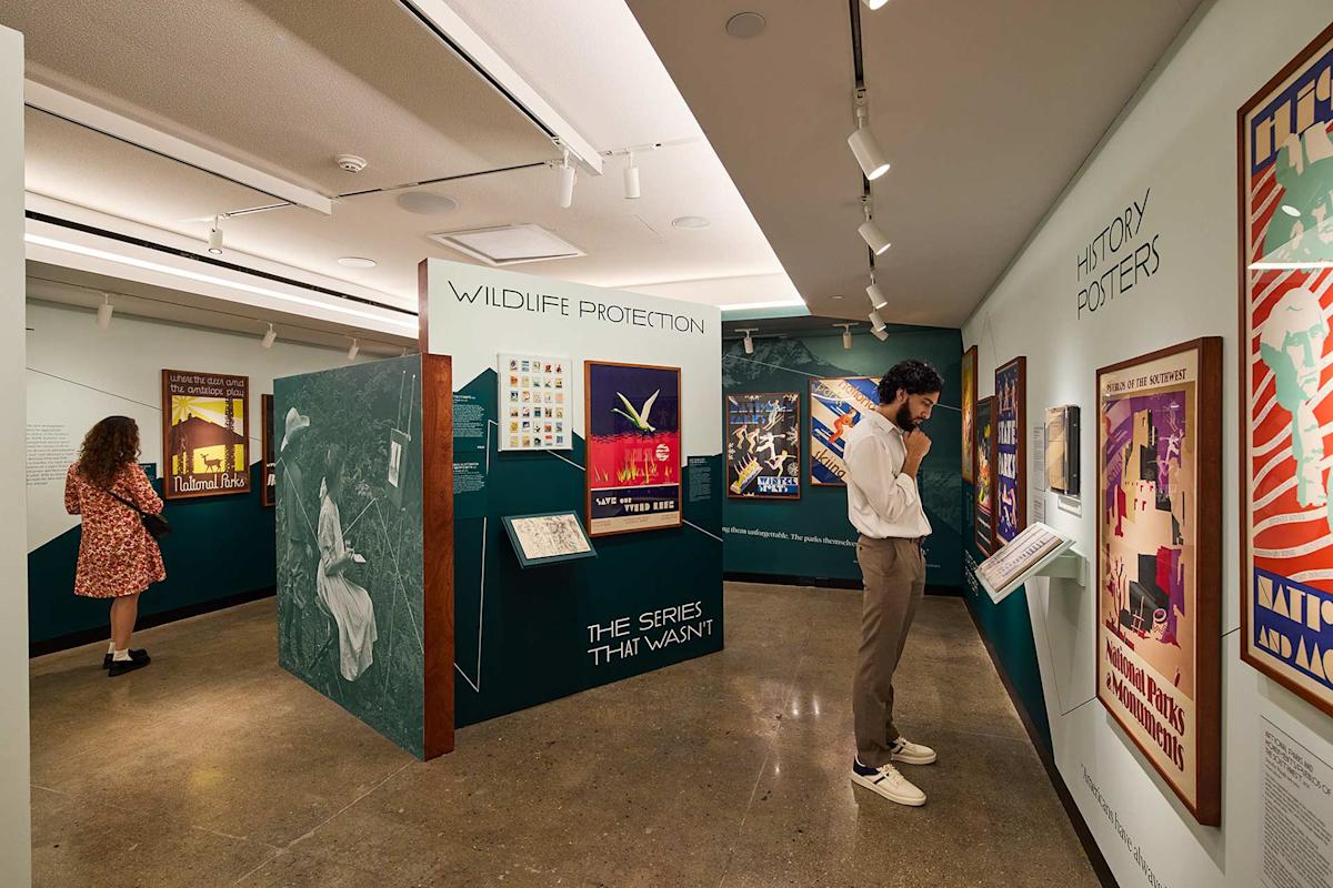 A museum exhibit features vintage wildlife protection and history posters. Two people observe displays on teal walls under bright lighting. The polished concrete floor reflects the vibrant, colorful posters.