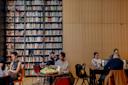 People studying and working on laptops at tables in a library. The room features a large wall filled with bookshelves and another wall with wooden paneling. Some individuals are reading while others are focused on their screens.
