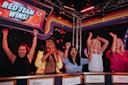 Five people stand behind a game show podium, cheering with raised arms as red confetti falls. A sign above reads “RED TEAM WINS!” The atmosphere is celebratory and joyful.