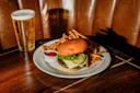 A plate with a cheeseburger topped with lettuce and pickles, a pile of French fries, and a small cup of ketchup, sits on a wooden table next to a glass of beer with a foamy head.