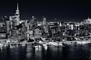 Black and white aerial view of the Manhattan skyline at night, featuring tall skyscrapers with illuminated windows reflecting on the water below.